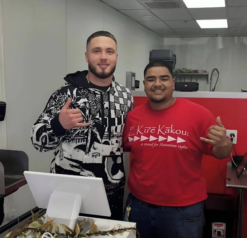 Two men smiling and making shaka signs in a restaurant kitchen with a POS system.