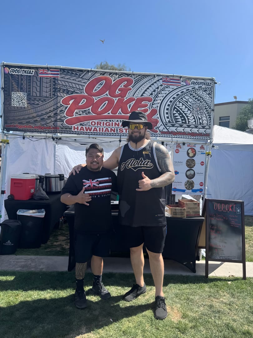 Two men stand smiling in front of an "OG Poke" food stall with a Hawaiian flag.